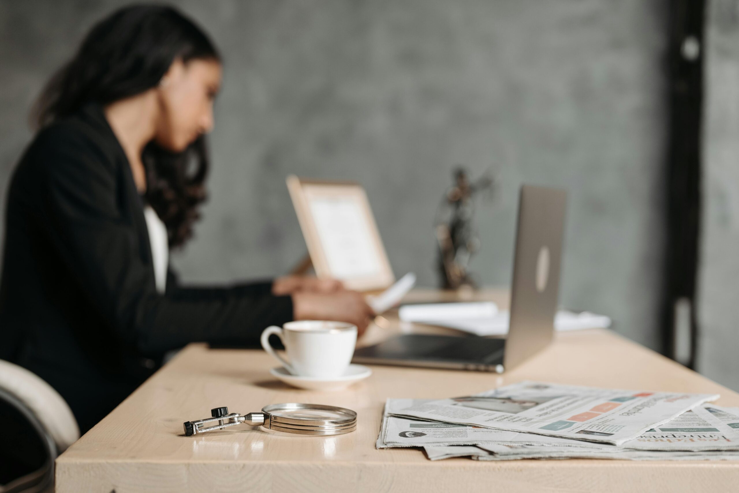 A woman in a business suit working at a desk with a laptop and papers in an office.